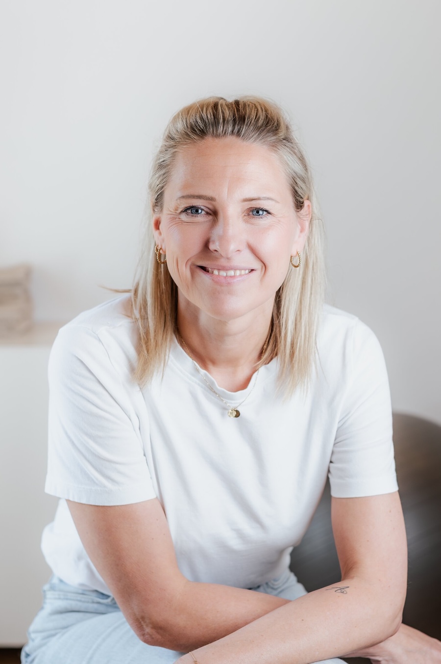 Porträt von Carina Brezina mit hellem Haar und weißem T-Shirt, lächelnd vor hellem Hintergrund.