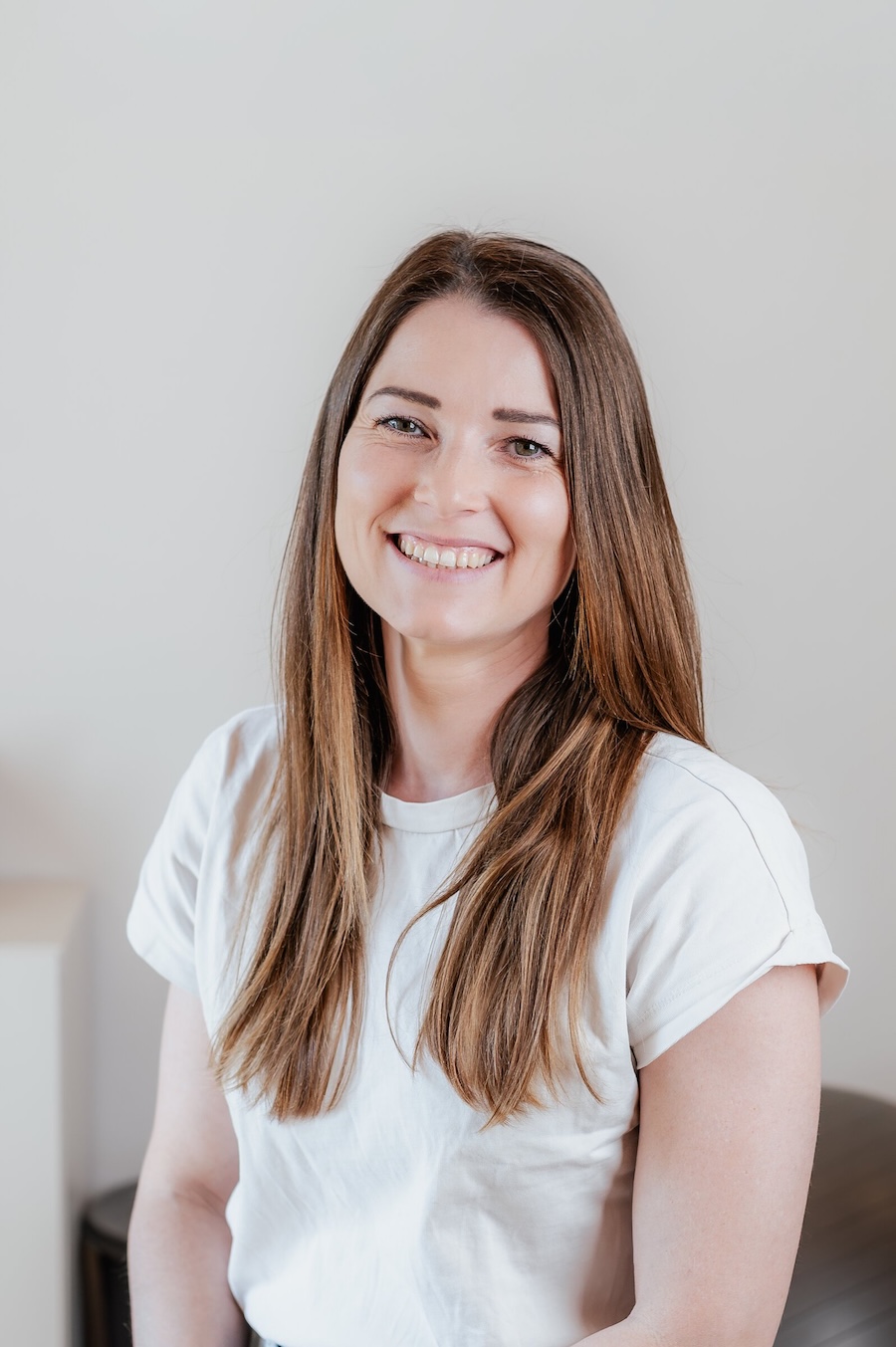 Porträt von Sabrina Scheucher mit langen braunen Haaren und weißem T-Shirt, lächelnd in einer hellen Umgebung.